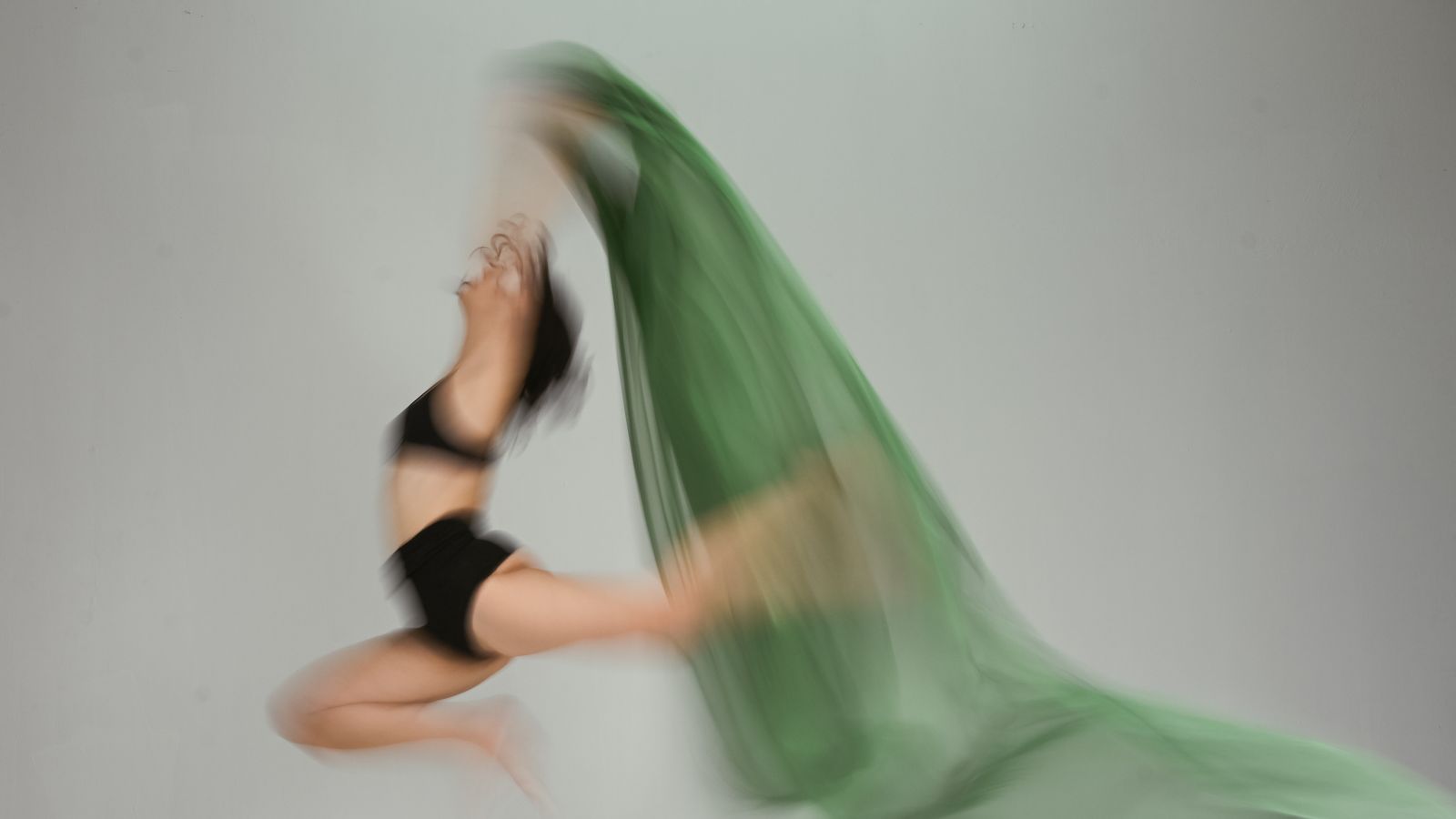 A dancer mid-leap with a green fabric trailing behind her in a motion blur against a light grey wall